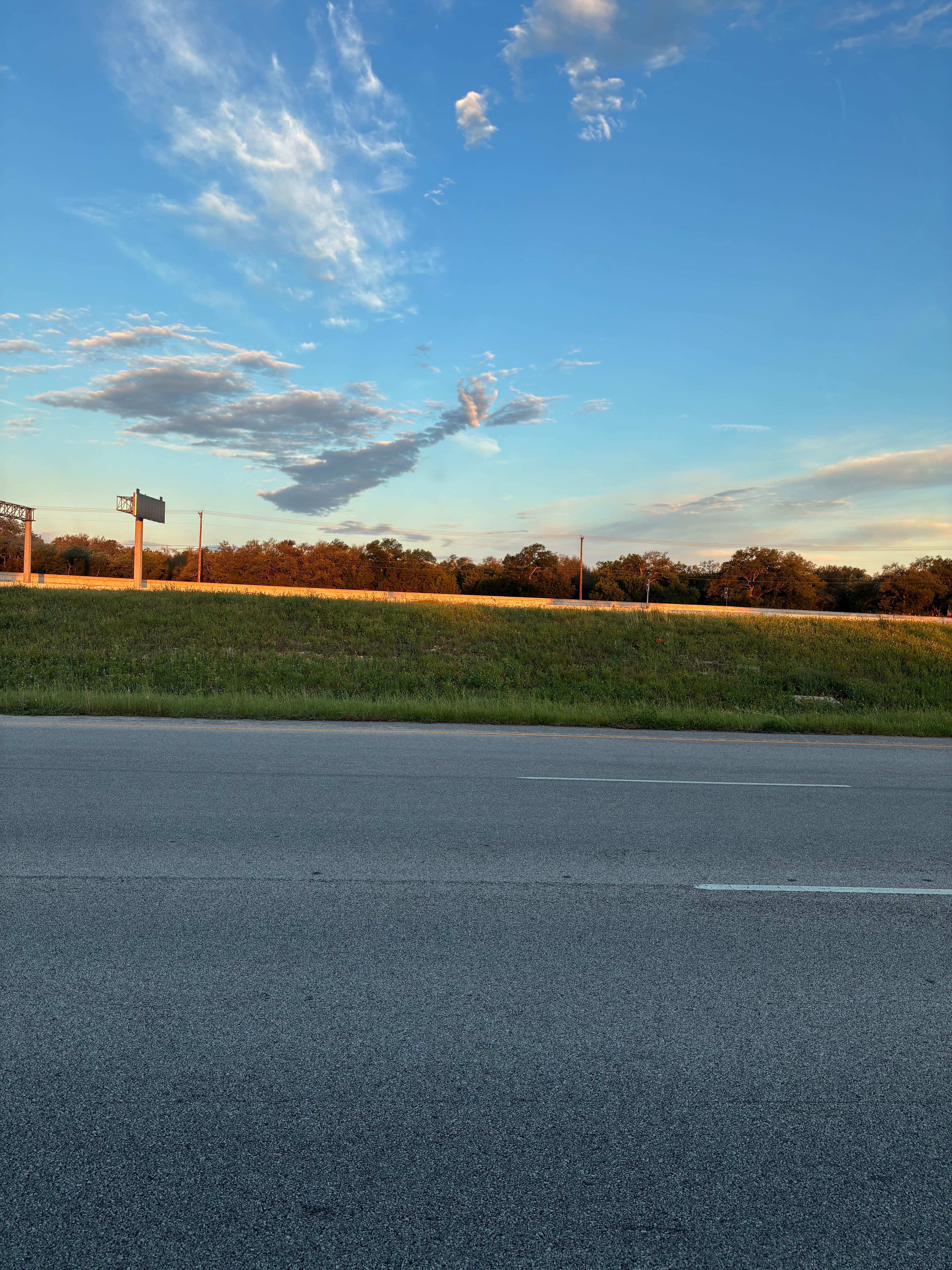 Asphalt highway beside a grassy field under a blue sky with clouds at golden hour.